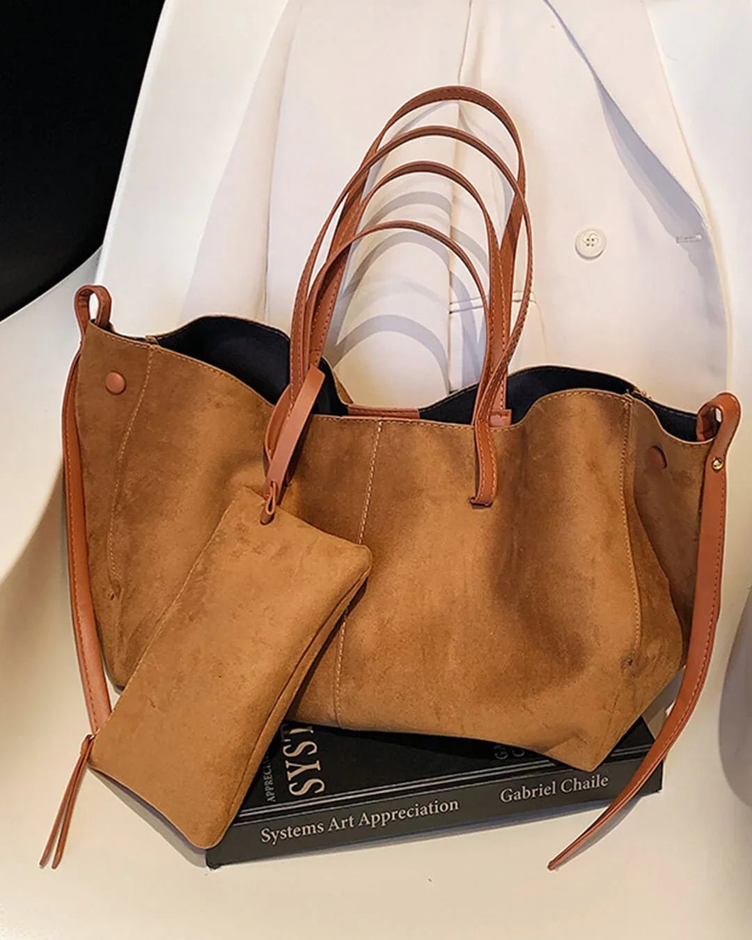 Brown suede tote bag with leather handles and matching pouch on a white chair